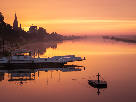 Aerial view of the Maas river at sunrise with the silhouette of Keverberg Castle and the church spire reflected in the calm water near a docked ferry boat in Kessel Limburg Netherlands.
