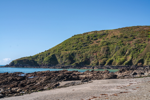View of the rugged coastline along the South West Coastal Path at Portnadler Bay with rocky outcrops sandy beach and green hills under a clear blue sky in Looe Cornwall United Kingdom. View of the rugged coastline along the South West Coastal Path at Portnadler Bay with rocky outcrops sandy beach and green hills under a clear blue sky in Looe Cornwall United Kingdom.