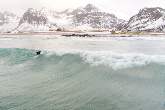 Aerial view of a surfer riding a wave with snow capped mountains in the background at Lofoten Nordland Norway.
