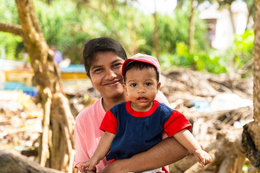 Sri Lanka 07 February 2026 View of a smiling woman holding a young child wearing a red cap in a lush tropical outdoor setting.
