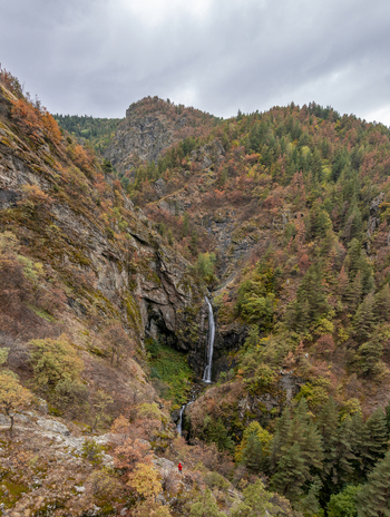 Aerial view of Ovcharenski Waterfall Goritsa flowing through a rocky mountain gorge surrounded by autumn foliage with a person standing at the base in Ovchartsi Kyustendil Province Bulgaria. Aerial view of Ovcharenski Waterfall Goritsa flowing through a rocky mountain gorge surrounded by autumn foliage with a person standing at the base in Ovchartsi Kyustendil Province Bulgaria.