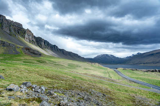 View of the Hvalfjordur fjord with steep rocky mountains green grassy slopes and a winding road under a dramatic cloudy sky in Iceland. View of the Hvalfjordur fjord with steep rocky mountains green grassy slopes and a winding road under a dramatic cloudy sky in Iceland.