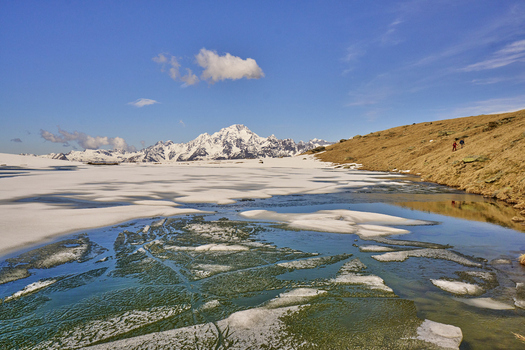 View of Valmalenco with a partially frozen lake snow covered mountain peaks and hikers on the grassy slope under a clear blue sky in Lombardy Italy. View of Valmalenco with a partially frozen lake snow covered mountain peaks and hikers on the grassy slope under a clear blue sky in Lombardy Italy.
