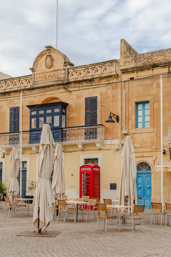 View of old buildings with blue doors a red telephone box and tables with umbrellas standing on the square Marsaxlokk Malta. View of old buildings with blue doors a red telephone box and tables with umbrellas standing on the square Marsaxlokk Malta.