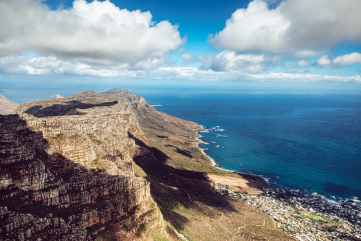 View of the rugged cliffs of Table Mountain overlooking the blue Atlantic Ocean and coastal suburbs under a cloudy sky in Cape Town Western Cape South Africa. View of the rugged cliffs of Table Mountain overlooking the blue Atlantic Ocean and coastal suburbs under a cloudy sky in Cape Town Western Cape South Africa.
