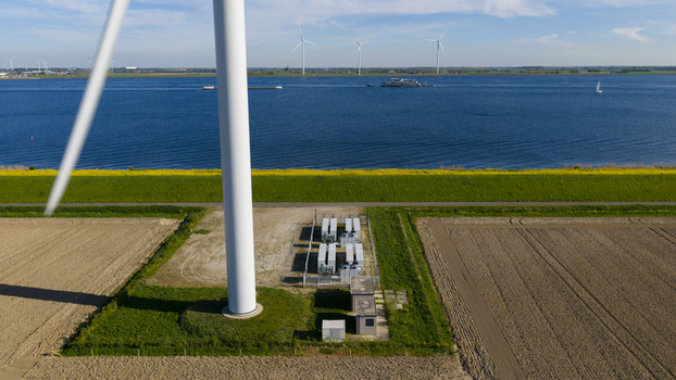 Aerial view of a large wind turbine base and electrical battery storage units next to agricultural fields and a blue waterway with ships in Ooltgensplaat Zuid Holland Netherlands. Aerial view of a large wind turbine base and electrical battery storage units next to agricultural fields and a blue waterway with ships in Ooltgensplaat Zuid Holland Netherlands.