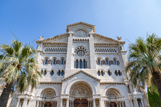 View of the Saint Nicholas Cathedral facade with ornate stone carvings a rose window and palm trees under a clear blue sky in Monaco City Monaco. View of the Saint Nicholas Cathedral facade with ornate stone carvings a rose window and palm trees under a clear blue sky in Monaco City Monaco.