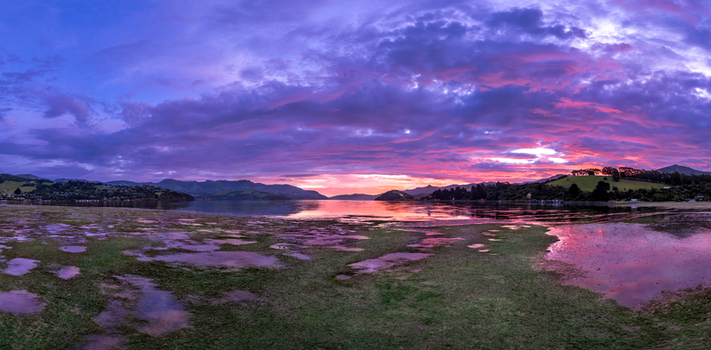 Aerial view of Duvauchelle bay at sunset with vibrant purple and pink clouds reflecting on calm water and exposed mudflats at low tide Duvauchelle Canterbury Region New Zealand. Aerial view of Duvauchelle bay at sunset with vibrant purple and pink clouds reflecting on calm water and exposed mudflats at low tide Duvauchelle Canterbury Region New Zealand.