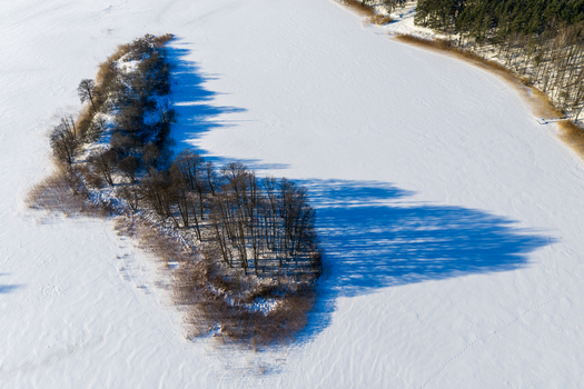 Aerial view of a small tree covered island in the middle of a frozen snow covered lake with long shadows and a winter forest shoreline Lithuania. Aerial view of a small tree covered island in the middle of a frozen snow covered lake with long shadows and a winter forest shoreline Lithuania.