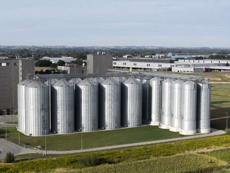 Aerial view of a large industrial grain silo complex with multiple metallic cylindrical storage tanks situated next to agricultural fields and industrial buildings in Poland. Aerial view of a large industrial grain silo complex with multiple metallic cylindrical storage tanks situated next to agricultural fields and industrial buildings in Poland.