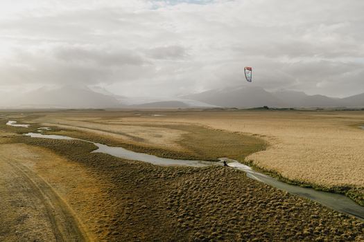 Aerial view of a kitesurfer gliding along a winding river through a vast golden landscape with distant mountains under a cloudy sky in Hofn Sveitarfelagid Hornaf Iceland.