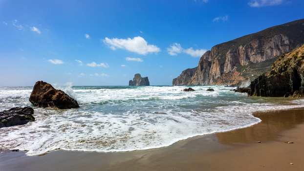View of Scoglio Pan di zucchero sea stack from the sandy shore of Masua beach with crashing waves and rugged cliffs under a blue sky Masua Sardegna Italy. View of Scoglio Pan di zucchero sea stack from the sandy shore of Masua beach with crashing waves and rugged cliffs under a blue sky Masua Sardegna Italy.