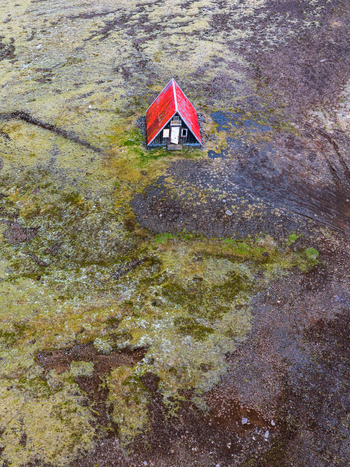 Aerial view of a small A frame cabin with a vibrant red roof situated in the middle of a vast moss covered volcanic landscape in Iceland. Aerial view of a small A frame cabin with a vibrant red roof situated in the middle of a vast moss covered volcanic landscape in Iceland.