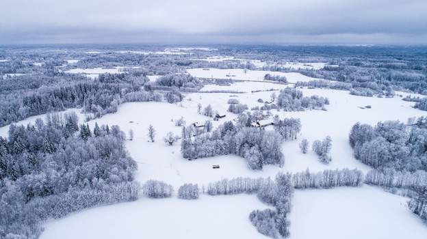 Aerial view of a snow covered rural landscape featuring dense forests open fields and a small cluster of farm buildings under an overcast winter sky Latvia. Aerial view of a snow covered rural landscape featuring dense forests open fields and a small cluster of farm buildings under an overcast winter sky Latvia.