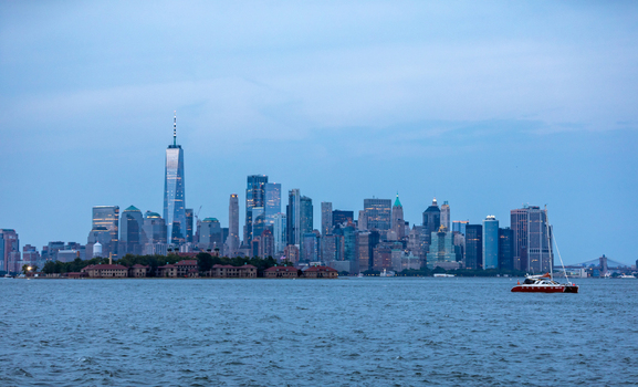 New York New York United States 08 July 2021 View of the Manhattan skyline and Ellis Island with a sailboat on the water under a clear blue sky. New York New York United States 08 July 2021 View of the Manhattan skyline and Ellis Island with a sailboat on the water under a clear blue sky.