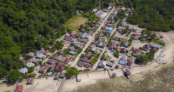Aerial view of a coastal village in Raja Ampat nestled between tropical forest and the sea featuring houses with metal roofs and wooden piers in Raja Ampat Southwest Papua Indonesia.