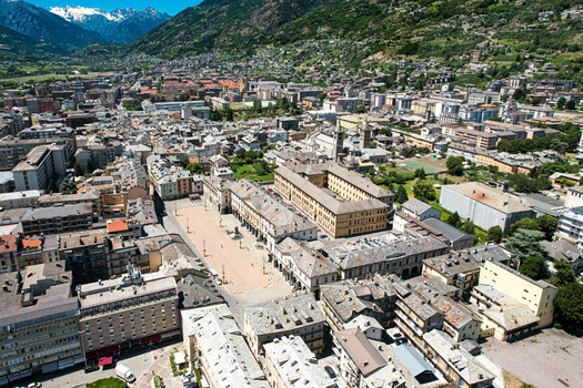 Aerial view of Piazza Chanoux and the historic center surrounded by traditional rooftops and the majestic snow capped Alps under a clear blue sky Aosta Aosta Valley Italy. Aerial view of Piazza Chanoux and the historic center surrounded by traditional rooftops and the majestic snow capped Alps under a clear blue sky Aosta Aosta Valley Italy.