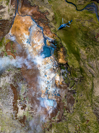 Aerial view of a steaming geothermal area featuring a vibrant blue pool mossy green terrain and a helicopter parked nearby in Iceland. Aerial view of a steaming geothermal area featuring a vibrant blue pool mossy green terrain and a helicopter parked nearby in Iceland.
