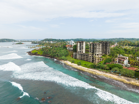 Aerial view of an unfinished concrete building on the coastline surrounded by palm trees and ocean waves under a cloudy sky in Mirissa Southern Province Sri Lanka. Aerial view of an unfinished concrete building on the coastline surrounded by palm trees and ocean waves under a cloudy sky in Mirissa Southern Province Sri Lanka.