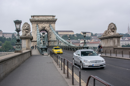 View of the Szechenyi Chain Bridge with its iconic stone lion statues a yellow taxi and a silver car driving across the bridge in Budapest Hungary.