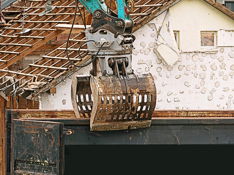 View of a heavy duty excavator with a demolition grapple tearing down a residential building structure with exposed roof rafters and damaged walls in Garching Bavaria Germany.