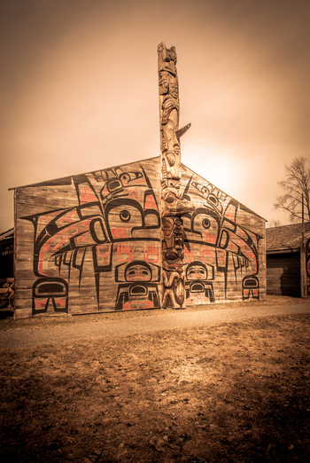 View of a traditional longhouse with intricate indigenous art and a tall carved totem pole at Ksan Historical Village in Ksan British Columbia Canada. View of a traditional longhouse with intricate indigenous art and a tall carved totem pole at Ksan Historical Village in Ksan British Columbia Canada.