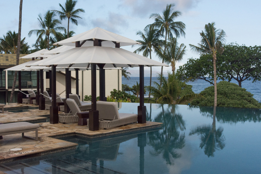 View of an infinity pool with lounge chairs and umbrellas overlooking the ocean surrounded by palm trees in Maui County Hawaii United States.