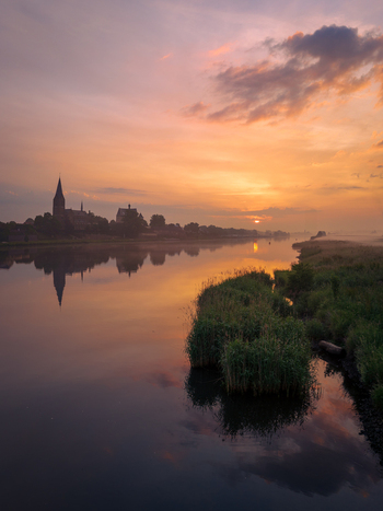 View of the historic church spire and village silhouette reflected in the calm Meuse river during a vibrant sunrise with morning mist Kessel Limburg Netherlands. View of the historic church spire and village silhouette reflected in the calm Meuse river during a vibrant sunrise with morning mist Kessel Limburg Netherlands.