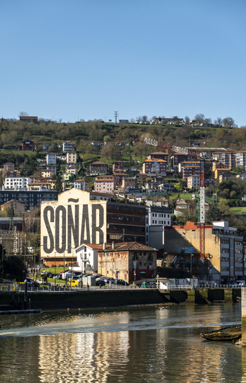 View of the prominent SONAR mural on a riverside building with residential houses on the hillside under a clear blue sky in Greater Bilbao Basque Country Spain. View of the prominent SONAR mural on a riverside building with residential houses on the hillside under a clear blue sky in Greater Bilbao Basque Country Spain.