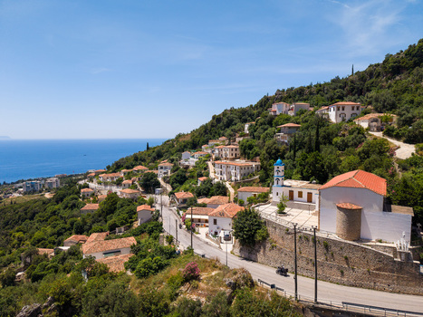 Aerial view of the coastal village featuring white houses with red tiled roofs and a church with a blue bell tower on a lush green hillside Dhermi Vlore County Albania.