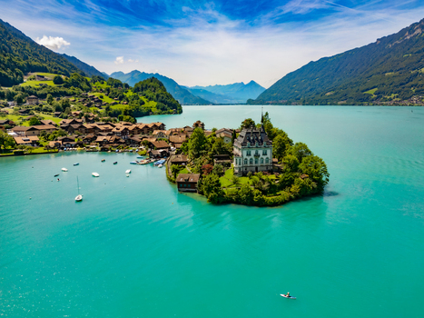 Aerial view of the turquoise lake embracing a quaint village and grand building nestled amidst verdant hills under a bright sky Iseltwald Canton of Bern Switzerland. Aerial view of the turquoise lake embracing a quaint village and grand building nestled amidst verdant hills under a bright sky Iseltwald Canton of Bern Switzerland.