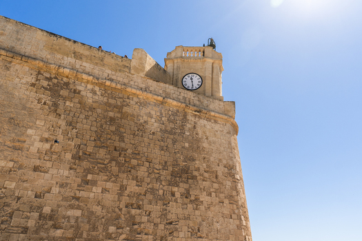 View of the massive limestone walls and a clock tower of a historic fortification under a clear blue sky in Zebbug Gozo Region Malta. View of the massive limestone walls and a clock tower of a historic fortification under a clear blue sky in Zebbug Gozo Region Malta.