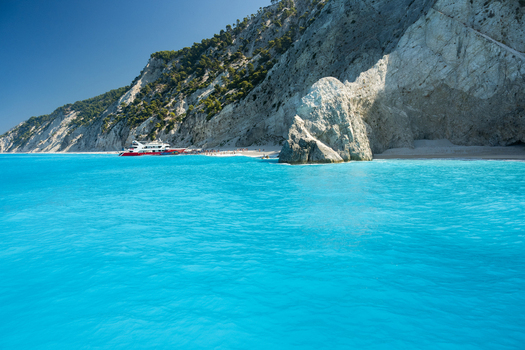 View of the turquoise waters of Porto Katsiki beach with a tour boat anchored near the white limestone cliffs under a clear blue sky in Lefkada Greece.