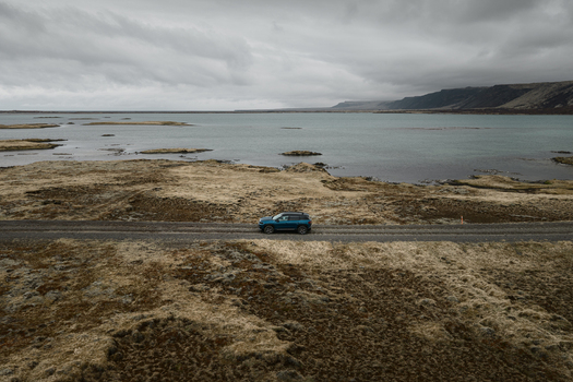 Aerial view of a blue SUV driving along a gravel road beside Hlidarvatn lake surrounded by mossy volcanic terrain and distant mountains in Sveitarfelagid Olfus Iceland. Aerial view of a blue SUV driving along a gravel road beside Hlidarvatn lake surrounded by mossy volcanic terrain and distant mountains in Sveitarfelagid Olfus Iceland.