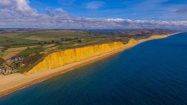 Aerial view of the golden sandstone cliffs stretching along the coastline with rolling green hills and deep blue sea West Bay England United Kingdom. Aerial view of the golden sandstone cliffs stretching along the coastline with rolling green hills and deep blue sea West Bay England United Kingdom.