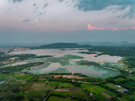 Aerial view of Fateh Sagar Lake surrounded by lush green fields and hills under a soft sunset sky in Udaipur Rajasthan India.