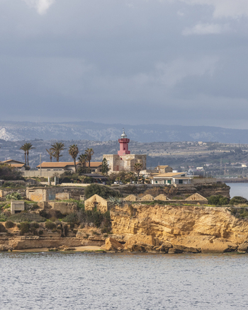 View of the red Capo Murro di Porco lighthouse on a rocky cliff overlooking the sea with palm trees and hills under a cloudy sky in Syracuse Sicily Italy.