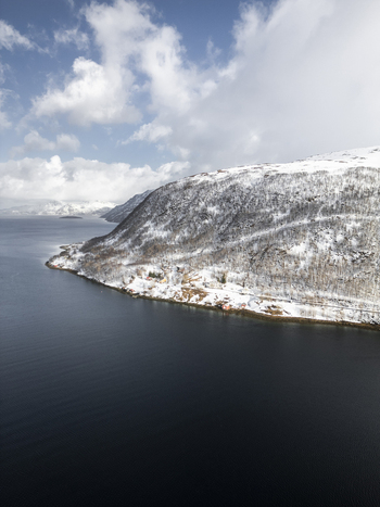 Aerial view of Grotfjord featuring snow covered mountain slopes scattered trees and small red houses along the dark blue water in Tromvik Troms Norway. Aerial view of Grotfjord featuring snow covered mountain slopes scattered trees and small red houses along the dark blue water in Tromvik Troms Norway.