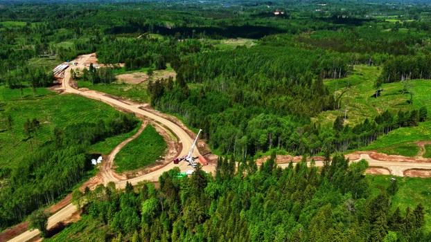 Aerial view of a wind turbine maintenance road winding through a dense green forest with construction equipment and a crane visible in Latvia. Aerial view of a wind turbine maintenance road winding through a dense green forest with construction equipment and a crane visible in Latvia.