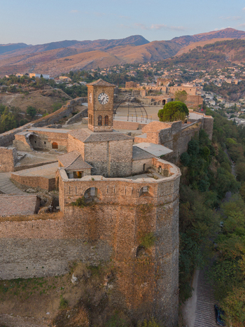 Aerial view of Gjirokaster Fortress with its clock tower stone walls and mountainous landscape under golden hour light in Gjirokaster Gjirokaster County Albania. Aerial view of Gjirokaster Fortress with its clock tower stone walls and mountainous landscape under golden hour light in Gjirokaster Gjirokaster County Albania.