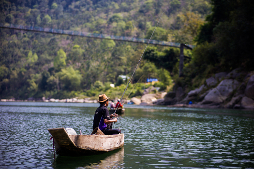 Meghalaya India 31 March 2023 View of a fisherman in a wooden boat casting a fishing line on a river with a suspension bridge in the background. Meghalaya India 31 March 2023 View of a fisherman in a wooden boat casting a fishing line on a river with a suspension bridge in the background.