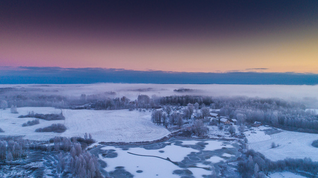 Aerial view of a frozen lake and snow covered fields surrounded by frost covered trees under a vibrant twilight sky in Latvia. Aerial view of a frozen lake and snow covered fields surrounded by frost covered trees under a vibrant twilight sky in Latvia.