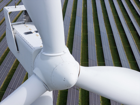 Aerial view of a wind turbine generator positioned within a large solar farm with rows of photovoltaic panels on green grass in Poland.