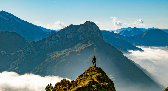 View of a person standing on a rocky peak in the Swiss Alps overlooking a vast mountain range with clouds in the valleys under a clear blue sky in Airolo Ticino Switzerland. View of a person standing on a rocky peak in the Swiss Alps overlooking a vast mountain range with clouds in the valleys under a clear blue sky in Airolo Ticino Switzerland.