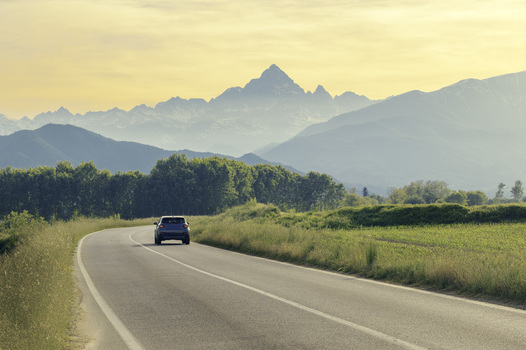 View of a car driving on a winding road towards the majestic Monviso mountain peak surrounded by green fields and trees under a soft golden sunset sky Oncino Piedmont Italy.