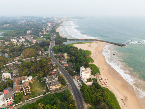 Aerial view of the coastal road running alongside a sandy beach with people umbrellas ocean waves and lush greenery in Galle Southern Province Sri Lanka.