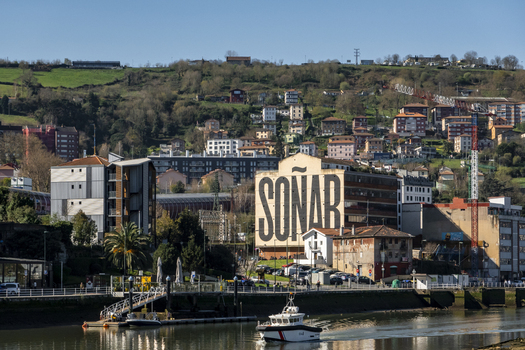 View of the Nervion river with a boat passing by the SONAR mural and hillside residential buildings under a clear sky in Greater Bilbao Basque Country Spain. View of the Nervion river with a boat passing by the SONAR mural and hillside residential buildings under a clear sky in Greater Bilbao Basque Country Spain.