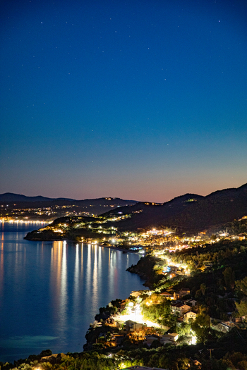 Aerial view of the illuminated coastline and town at night with lights reflecting on the calm sea under a starry sky in Corfu Ionian Islands Greece. Aerial view of the illuminated coastline and town at night with lights reflecting on the calm sea under a starry sky in Corfu Ionian Islands Greece.
