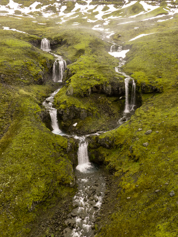 Aerial view of a series of cascading waterfalls flowing down a vibrant green mossy hillside with patches of snow in the Eastern Region Iceland. Aerial view of a series of cascading waterfalls flowing down a vibrant green mossy hillside with patches of snow in the Eastern Region Iceland.