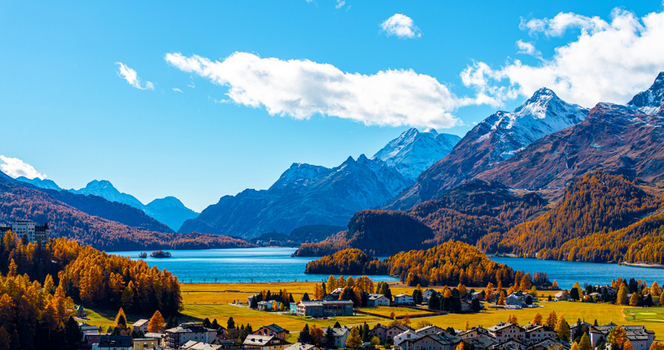 Aerial view of Lake Sils with golden autumn larch forests snow capped mountains and a small village in the foreground under a clear blue sky in Maloja District Grisons Switzerland.